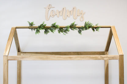 A cozy bedroom with a neatly made bed is adorned with a wooden name sign hanging above the headboard. The sign has a rustic finish, adding a personalized and charming touch to the space.